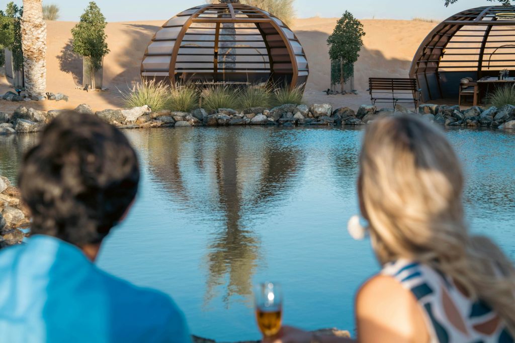 돔 구조물과 나무를 배경으로 연못이 내려다보이는 두 사람. 한 사람이 잔을 들고 있습니다.