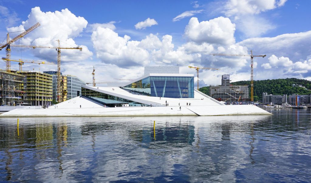 바닷가 옆에 경사진 지붕, 흩어져 걷는 사람들, 배경에 크레인, 흐린 하늘이 있는 현대적인 유리 건물.