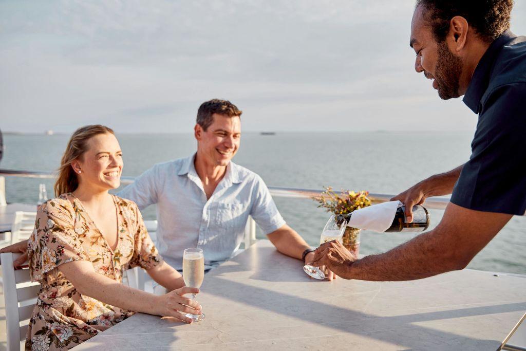 보트 위의 야외 테이블에서 서버가 유리잔에 샴페인을 부으며 미소를 짓고 있는 커플의 모습은 바다를 배경으로 펼쳐집니다.