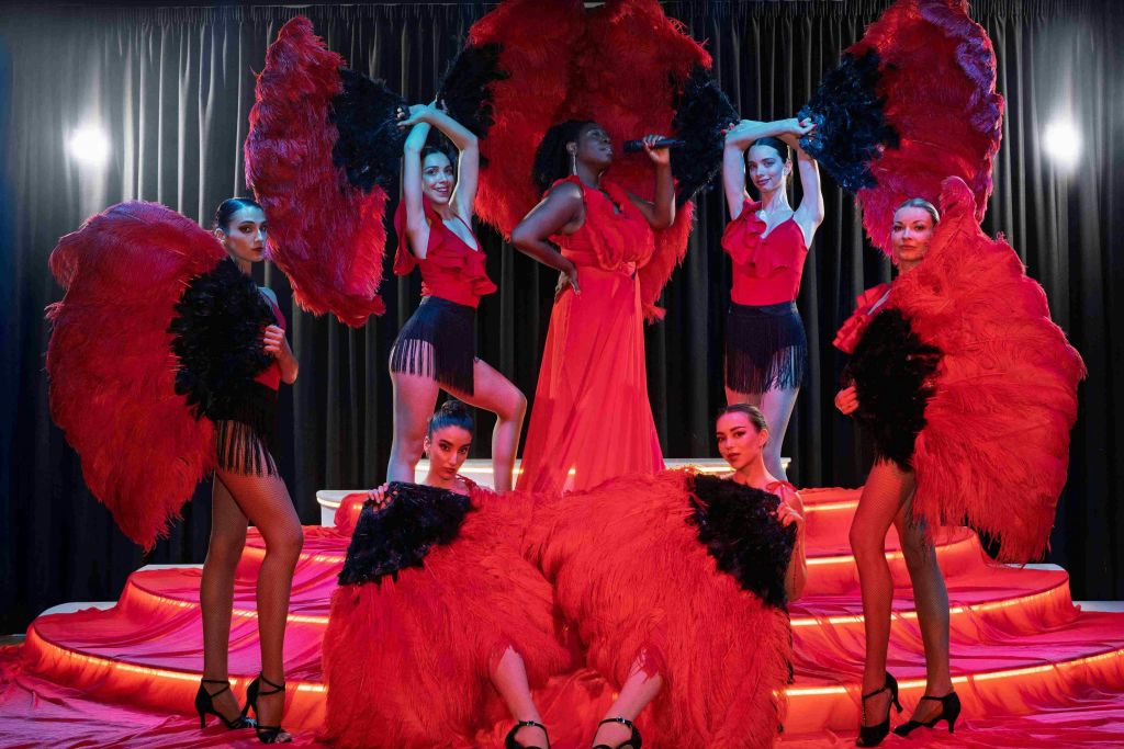 빨간색 드레스를 입은 연주자가 불이 켜진 무대에서 커다란 빨간색과 검은색 깃털 팬을 들고 있는 댄서들에게 둘러싸여 노래를 부릅니다.