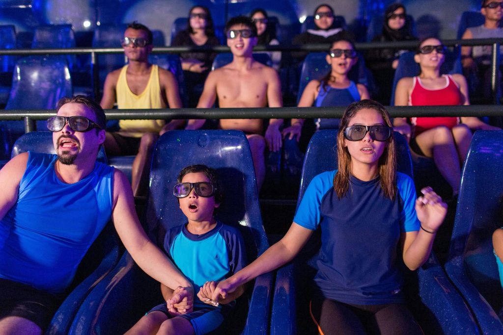 3D 안경을 쓰고 극장식 라이드에 앉은 사람들 중 일부는 몰입형 쇼를 경험하면서 손을 잡고 있습니다.