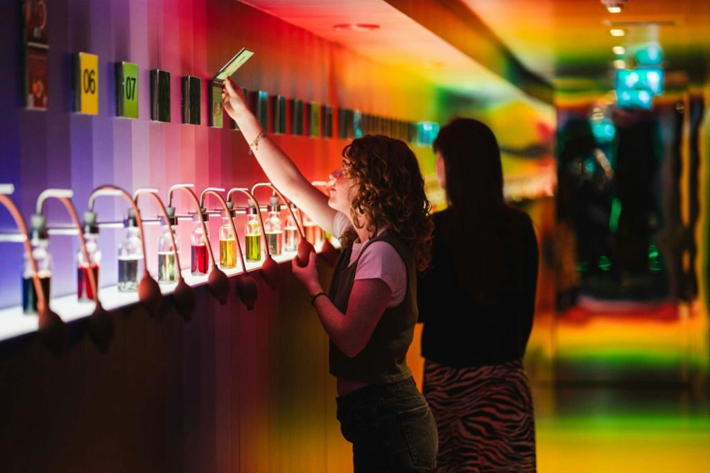 한 사람이 아래 작은 병이 놓인 무지개빛 벽에 카드를 놓으려고 손을 뻗고, 다른 한 사람이 근처에 서 있습니다.