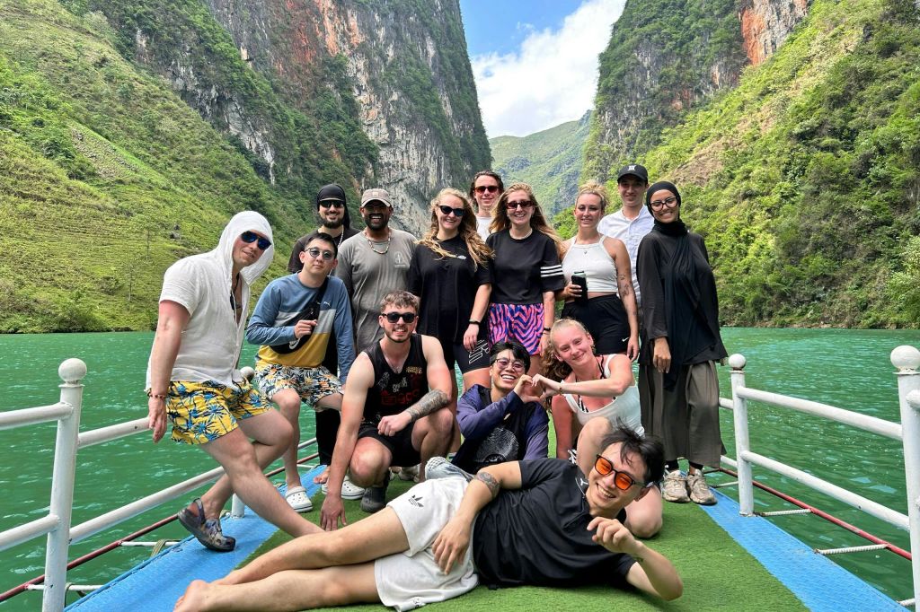 푸른 하늘이 펼쳐진 경치 좋고 울창한 산악 지형에 둘러싸인 한 무리의 사람들이 보트에서 함께 웃으며 포즈를 취하고 있습니다.