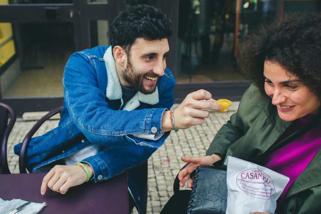한 남성이 바둑판식 포장 도로에서 야외에서 빨간색 문구가 적힌 흰 종이 봉투를 들고 앉아 있는 여성에게 음식을 제공하며 미소를 짓고 있습니다.