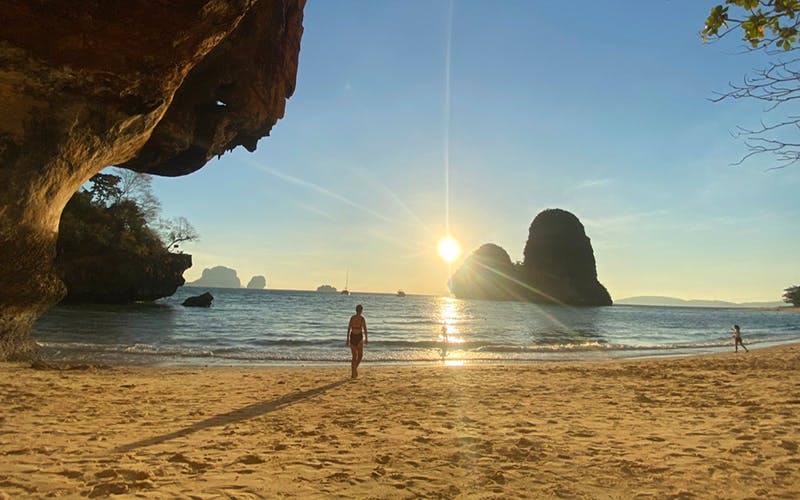 수영복을 입은 사람은 커다란 암석층과 석양을 배경으로 바다를 향해 모래사장을 거닐고 있습니다.