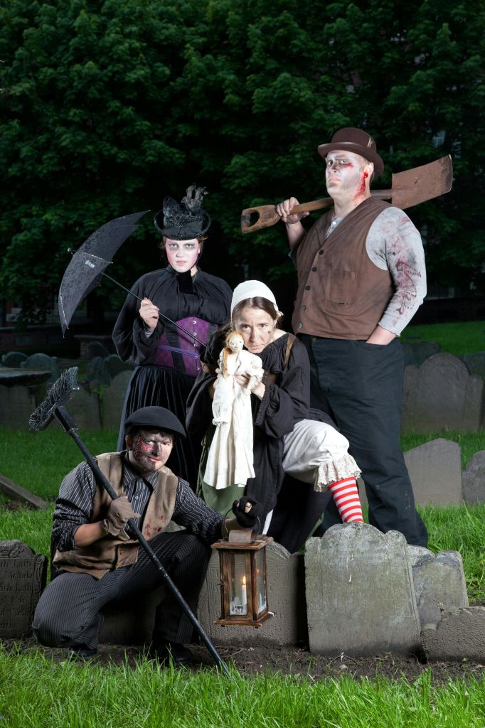 메이크업이 돋보이는 빅토리아 시대 스타일의 의상을 입은 네 명이 묘지에서 포즈를 취하고 있습니다. 한 명은 삽을 들고 다른 한 명은 인형을 들고 있습니다.