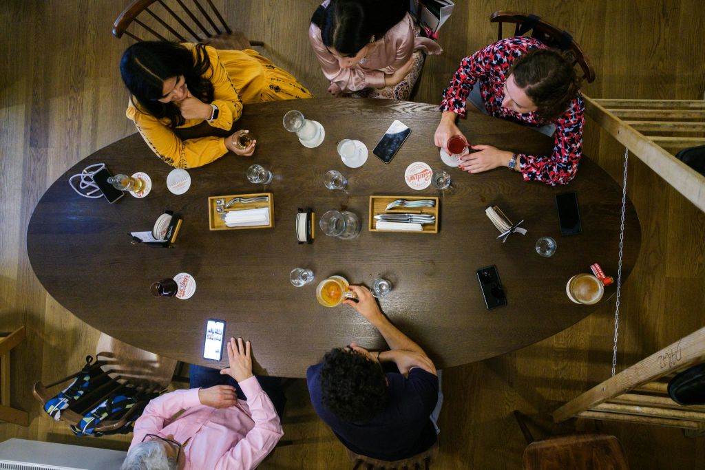 5명이 나무 테이블에 둘러앉아 음료, 전화기, 푸드트레이를 즐기고 있습니다. 위에서 본 모습.