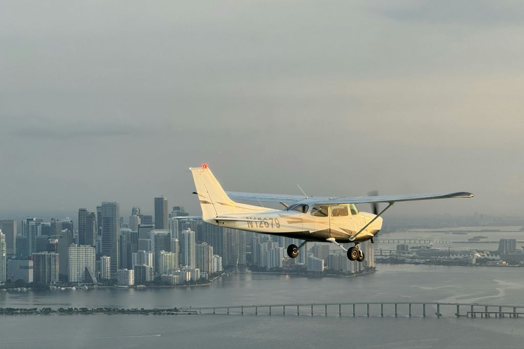 높은 건물이 있는 도시 풍경 위로 날아다니는 작은 비행기와 흐린 하늘 아래 물 위로 다리가 있습니다.