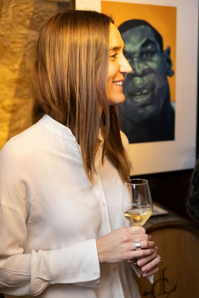 한 사람이 벽에 걸린 초상화 옆에 서서 음료를 들고 미소를 짓고 있습니다. 배경에는 따뜻한 조명과 질감의 벽이 있습니다.