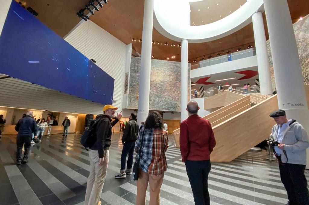 커다란 벽 디스플레이, 나무 계단, 자연 조명과 인공 조명이 어우러진 현대적인 복층 아트리움에 서 있는 사람들.