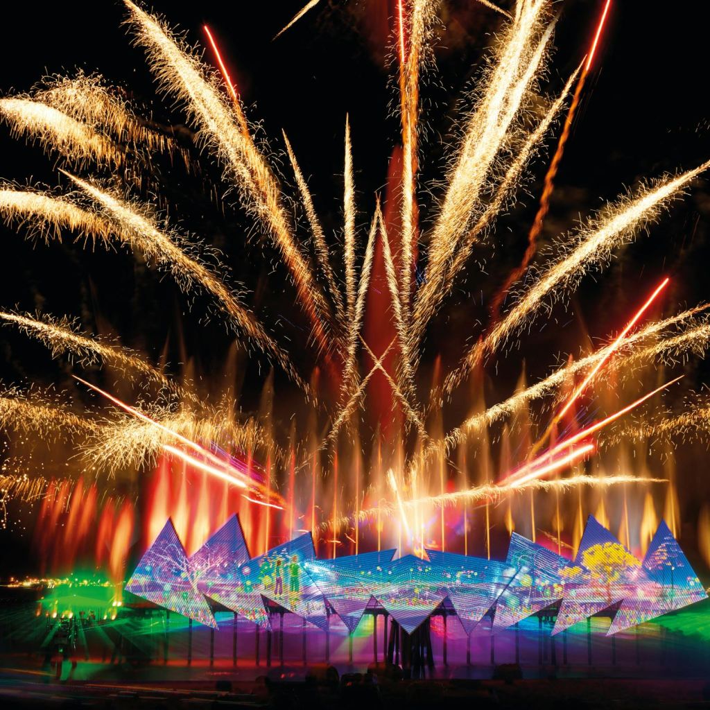 화려한 불꽃놀이가 활기찬 조명과 패턴으로 빛나는 기하학적 무대 위 밤하늘에서 펼쳐집니다.