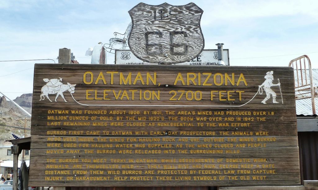애리조나주 오트만 (Oatman) 의 나무 표지판, 해발 2,700피트, 마을의 역사적인 광산 배경과 야생 버로에 대한 설명이 담긴 텍스트.