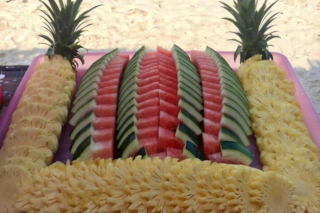 얇게 썬 수박과 파인애플이 테이블에 깔끔하게 배열되어 있고, 모서리에 전체 파인애플이 모래 배경에 표시됩니다.