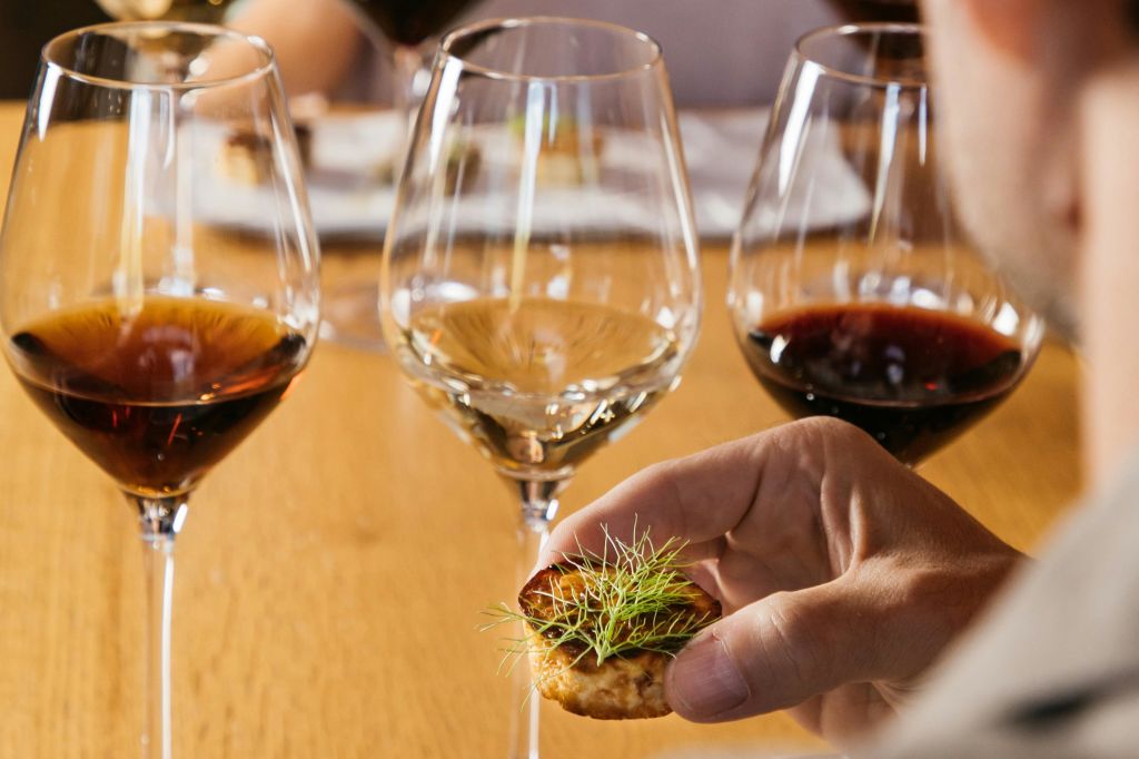 나무 테이블 위에 레드 와인 잔으로 둘러싸인 녹색의 작은 애피타이저를 들고 있는 손의 클로즈업.