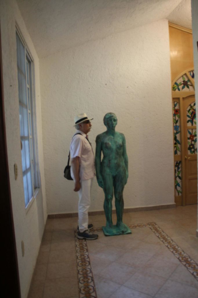 흰색 벽과 스테인드글라스 문이 있는 방의 녹색 청동 동상 옆에 흰색 복장을 한 남성이 서 있습니다.