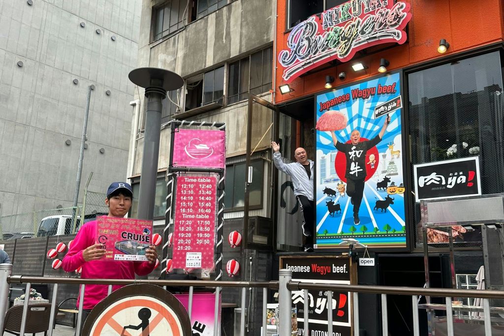 분홍색 옷을 입은 사람이 스케줄 보드 근처에서 표지판을 들고 있으며, 다른 사람이 근처 건물 창문에서 손을 흔들고 있습니다.