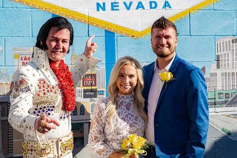 '네바다' 라고 쓰여진 벽화 앞에 세 사람이 서 있습니다. 왼쪽의 남자는 엘비스 의상을 입고 있습니다. 옆에 있던 부부가 꽃을 들고 있다.