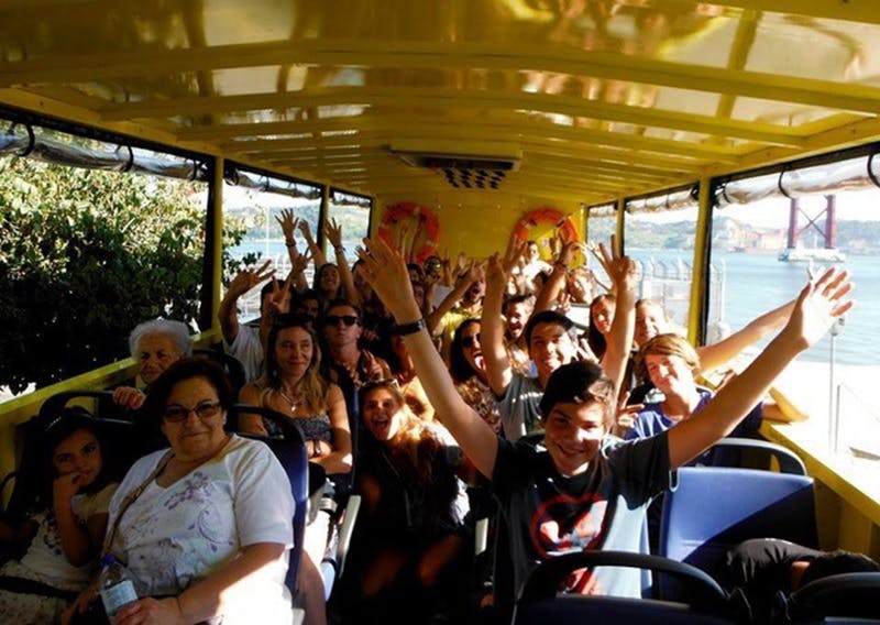 다양한 연령대의 사람들이 웃으며 손을 들고 열린 창문이 있는 노란색 투어 버스 안에 앉아 있습니다.