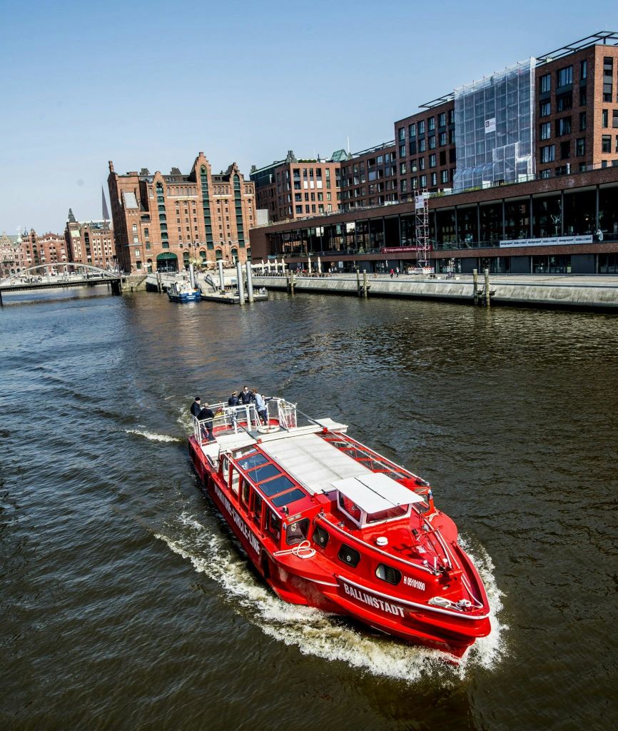 맑고 푸른 하늘 아래 유서 깊은 벽돌 건물과 현대적인 건축물이 즐비한 강을 따라 붉은 보트가 항해하고 있습니다.