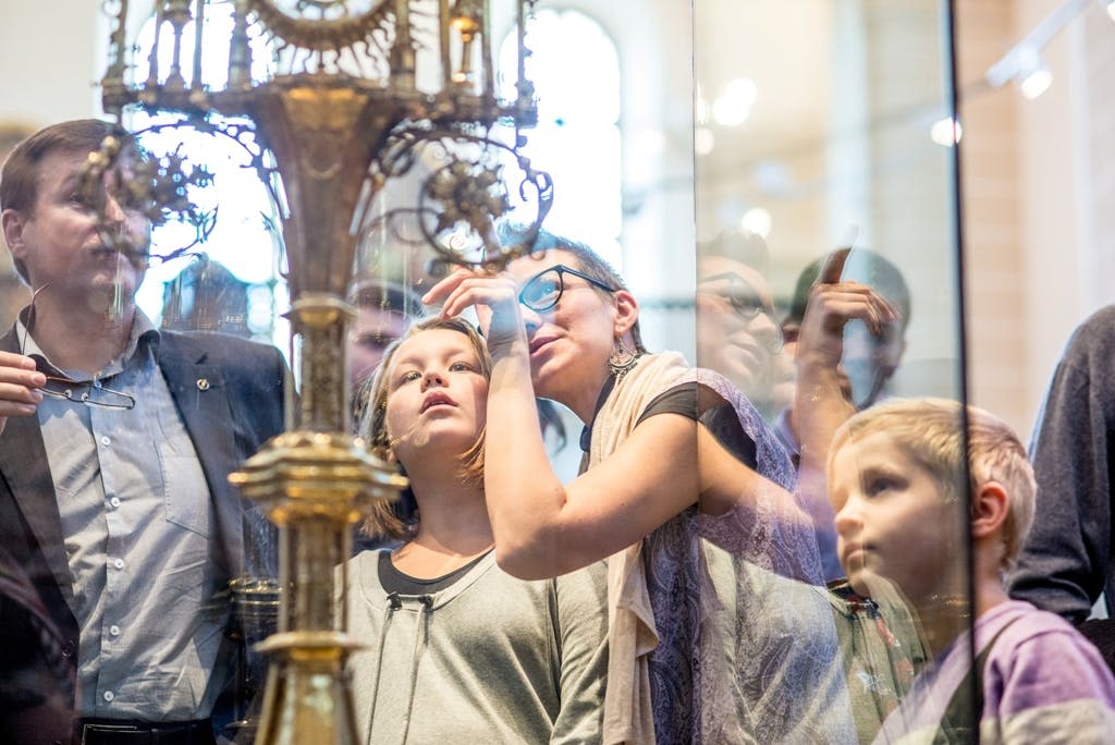 어린이와 성인을 포함한 한 무리의 사람들이 박물관에서 유리 뒤의 복잡한 화려한 물체를 면밀히 관찰합니다.