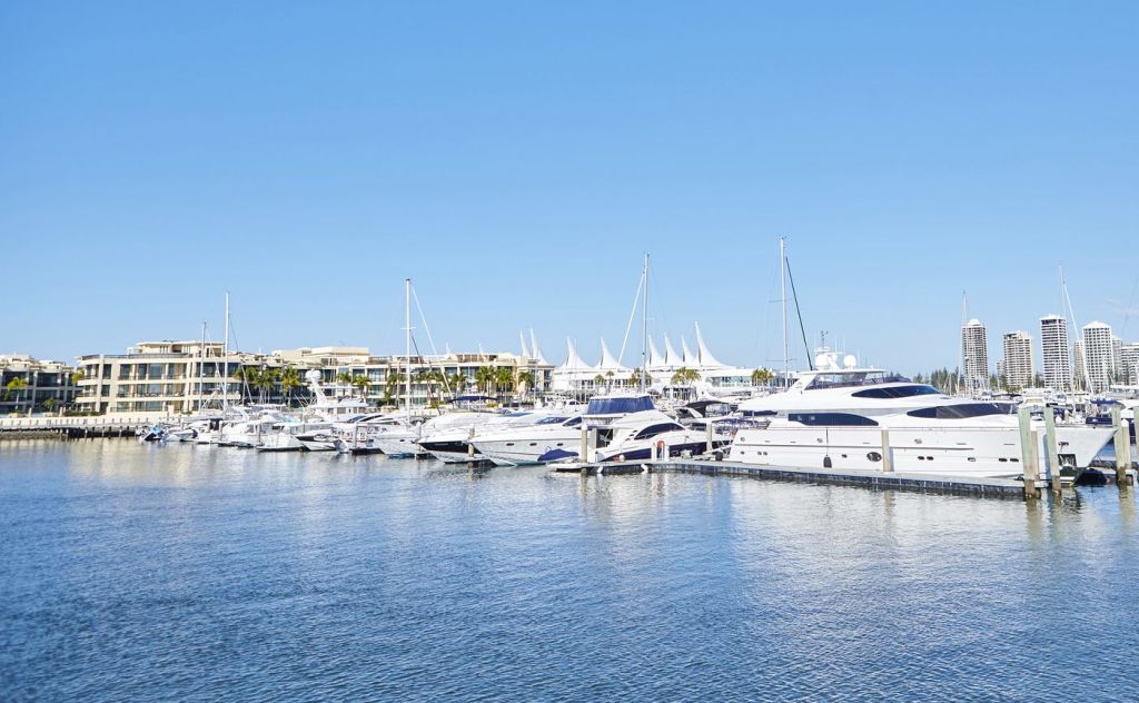 바닷가 옆에는 여러 대의 요트가 정박해 있는 정박지가 있습니다. 배경에는 머리 위로 푸른 하늘이 보이는 건물들이 있습니다.