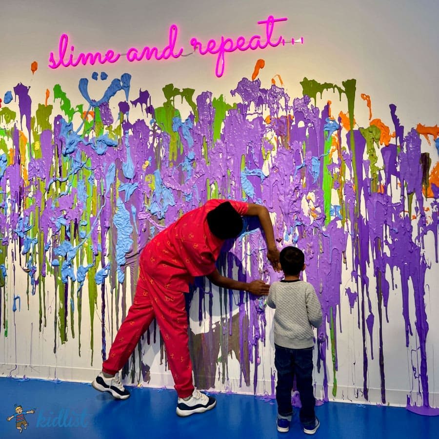 어른과 어린이는 분홍색 네온 불빛에 "점액질과 반복" 이라고 쓰인 표지판 아래 벽에 화려한 점액을 바르고 놀고 있습니다.