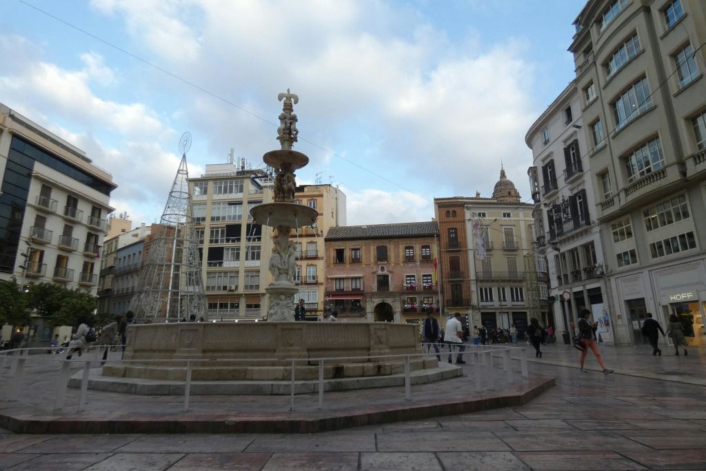 중앙 분수가 있는 마을 광장, 발코니가 있는 주변 건물, 산책하는 사람들. 금속 프레임워크 트리 구조가 있습니다.