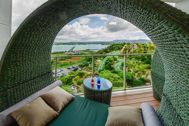 울창한 녹음이 내려다보이는 고리버들 캐노피 데이베드, 워터파크, 부분적으로 흐린 하늘 아래 호수에서 바라보는 전망. 테이블 위에 음료 두 잔.