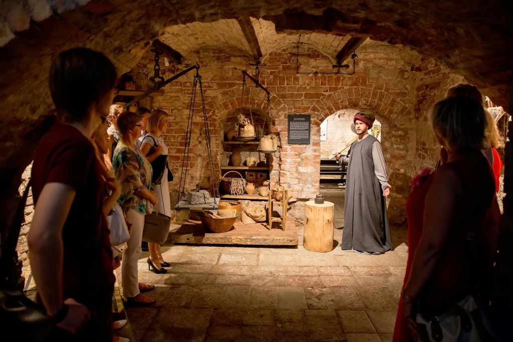 중세 복장을 한 가이드가 역사적 유물이 있는 어두운 조명, 아치형 벽돌 지하실에서 한 무리의 관광객과 이야기를 나눕니다.