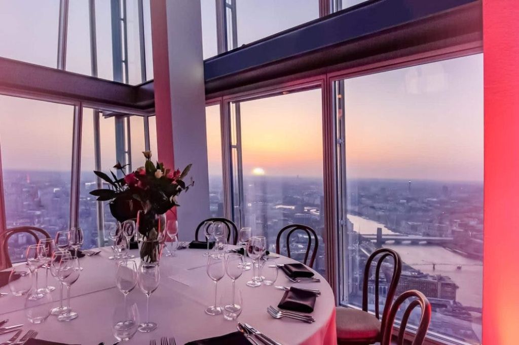 석양의 도시 풍경을 감상할 수 있는 커다란 창문 근처의 우아한 식사 공간. 꽃무늬 센터피스, 와인잔, 식기류가 놓인 테이블.