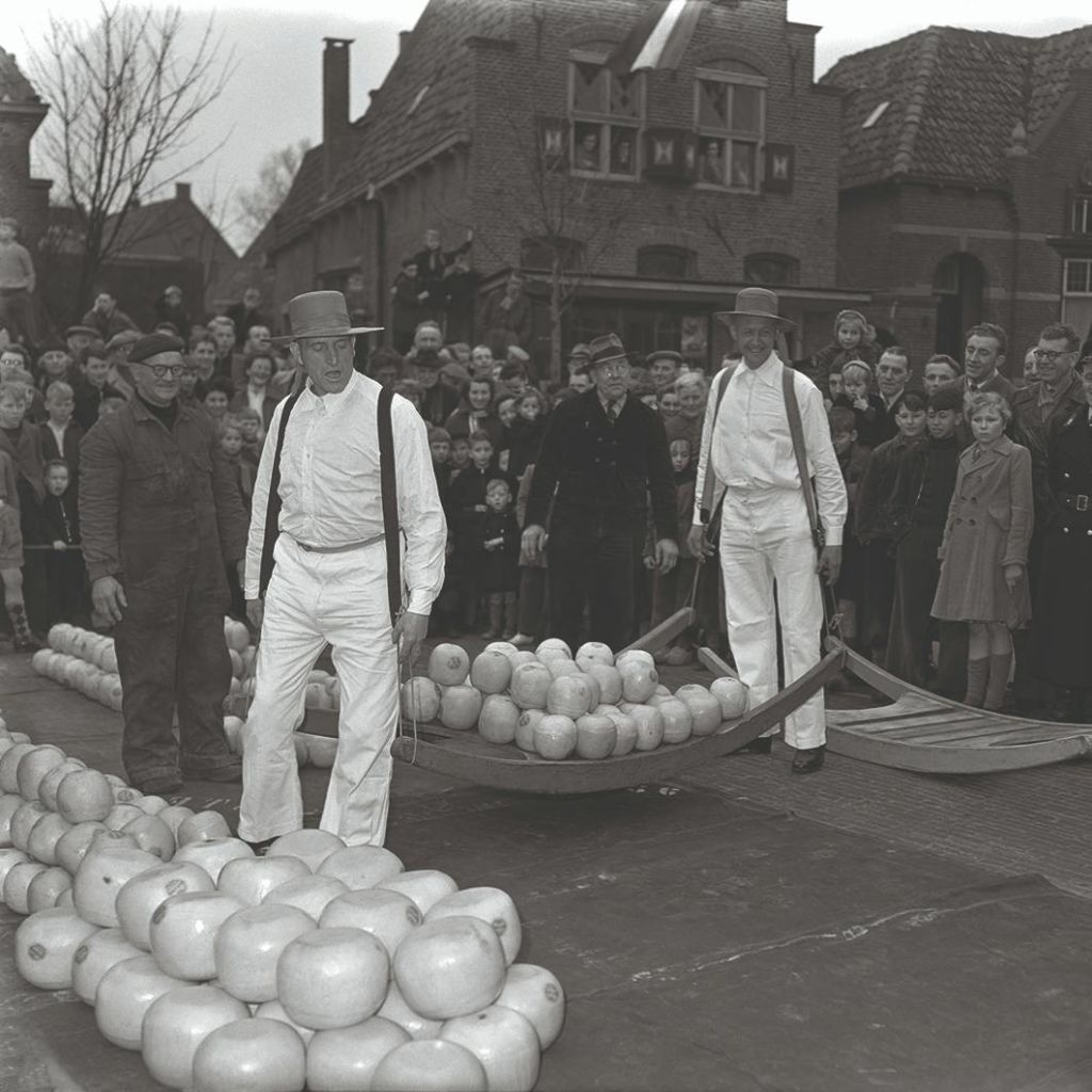 빈티지 거리에서 전통 복장을 한 남성들이 군중들 앞에서 치즈를 가득 담은 배로우를 들고 있습니다.