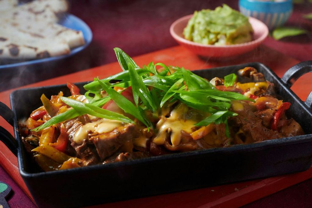 녹은 치즈와 파를 얹은 지글지글한 소고기 파히타, 과카몰리와 토르티야가 배경으로 보입니다.