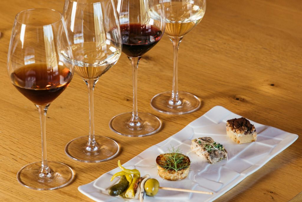 나무 테이블 위에 다양한 애피타이저가 놓인 흰색 직사각형 접시 옆에는 서로 다른 와인이 가득 담긴 와인 잔 네 개가 놓여 있습니다.