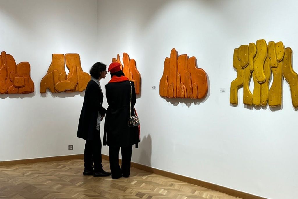 나무 마루가 깔린 조명이 밝은 갤러리에서 주황색과 노란색의 추상적인 벽 조형물을 감상하는 두 사람.