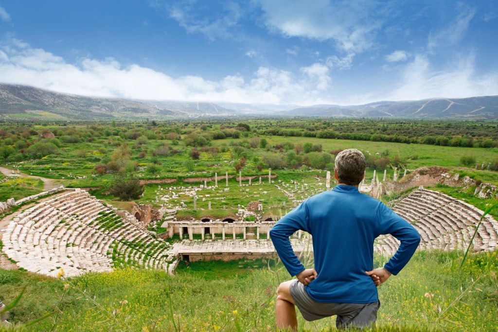푸른 셔츠를 입은 한 사람이 푸른 들판과 흐린 하늘로 둘러싸인 기둥과 석조 구조물이 있는 고대 유적을 내려다보고 있습니다.