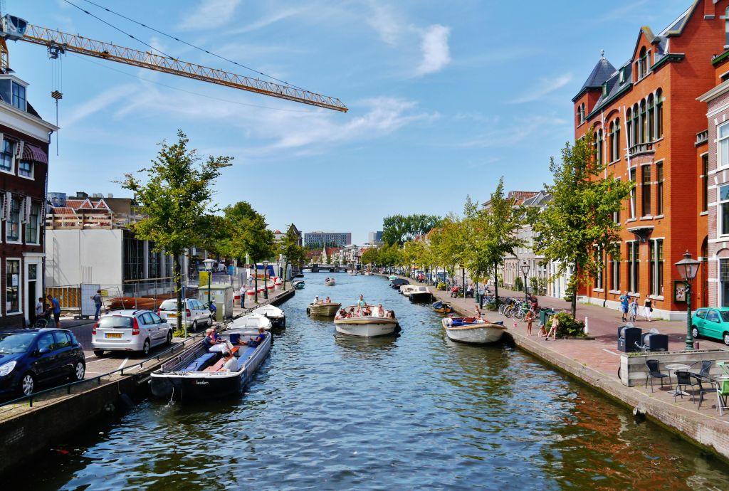 사람들은 화창한 날 나무, 주차된 차, 건물이 늘어선 운하를 헤엄치는 보트에서 휴식을 취합니다. 머리 위에는 크레인이 있습니다.