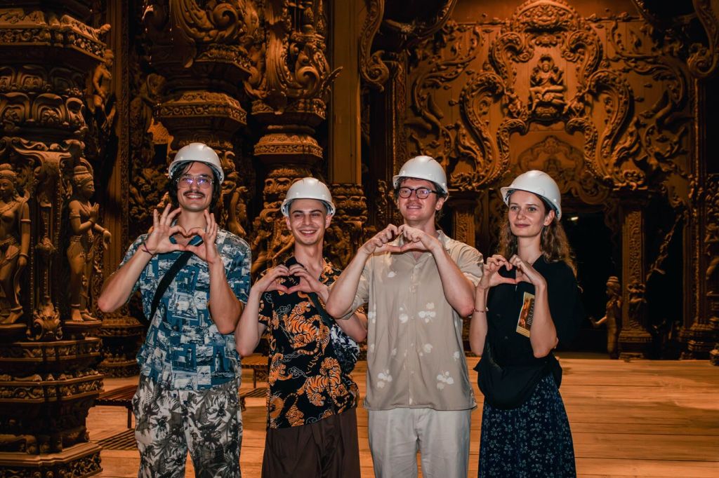 안전모와 캐주얼한 옷을 입은 네 명의 사람이 화려한 조각된 나무 배경에 손을 대고 하트 모양을 하고 있습니다.