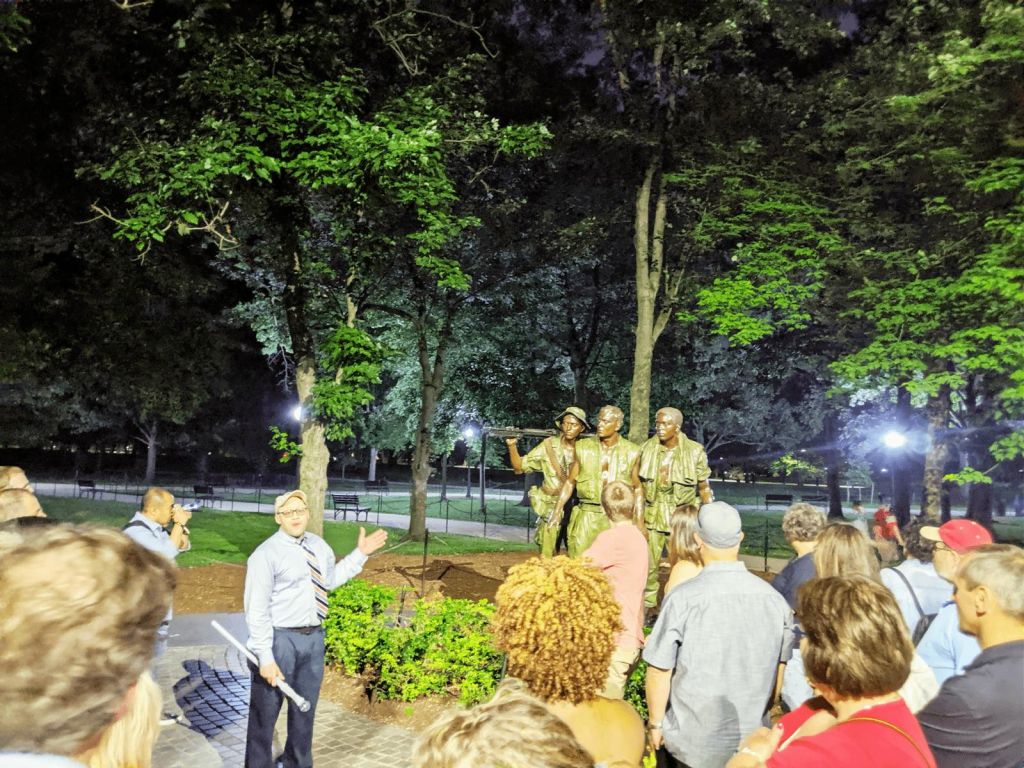 한 무리의 사람들이 밤에 공원에서 네 명의 군인 동상 앞에서 말하는 한 남자의 말을 듣고 있습니다.