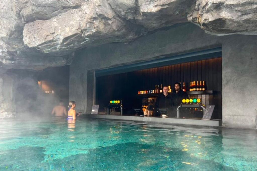바위 지층에 자리한 스윔업 바 뒤에 두 명의 바텐더가 서 있으며, 전경에 수영장이 있습니다. 물병과 수도꼭지가 보입니다.