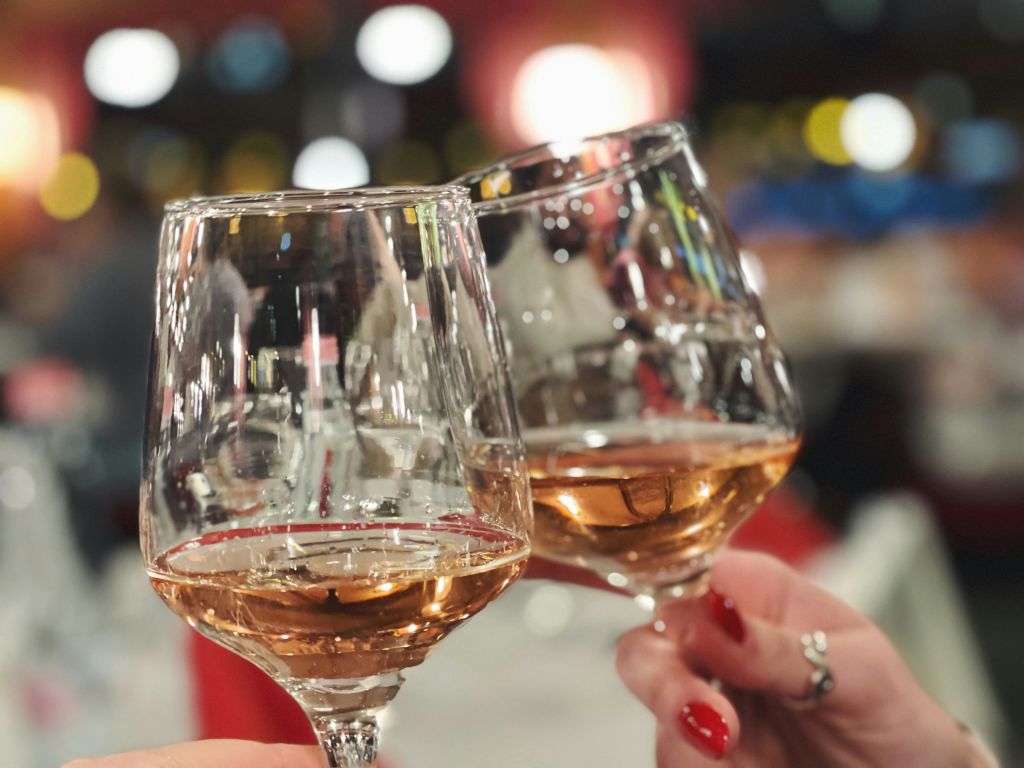 빨간색으로 칠해진 손톱으로 손을 잡은 두 개의 와인 잔이 화려한 불빛이 흐릿한 배경을 배경으로 토스트에 찰칵찰칵 들어옵니다.