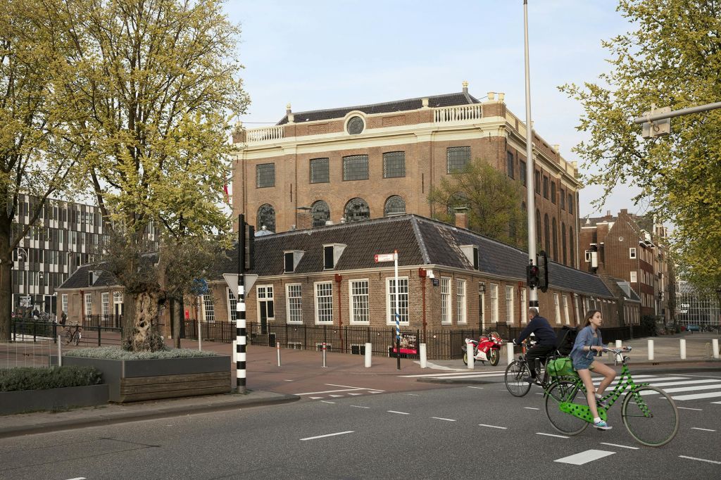 자전거를 타는 사람들, 아치형 창문이 있는 커다란 벽돌 건물, 나무로 둘러싸인 작은 벽돌 건물이 있는 거리 풍경.