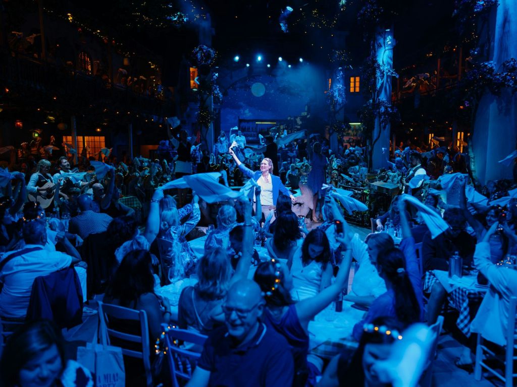 냅킨을 흔드는 사람들, 화려한 조명, 공연자가 중앙에 서 있는 활기찬 실내 행사입니다.