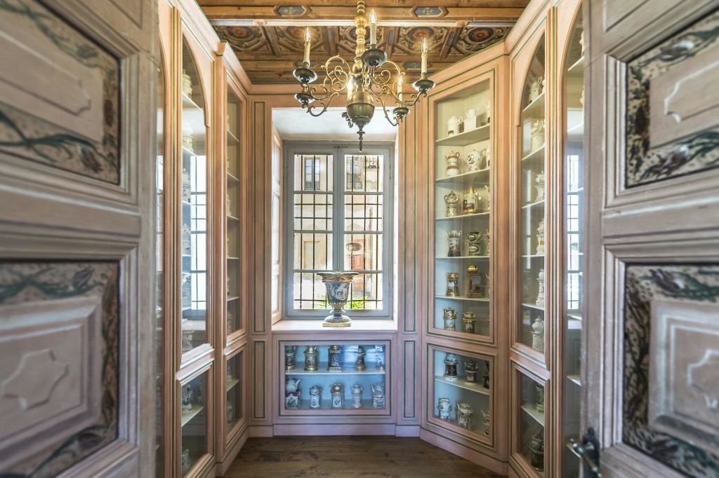 다양한 항아리를 전시한 나무 수납장과 중앙 창문, 장식된 천장에 매달린 샹들리에가 있는 작고 화려한 객실입니다.