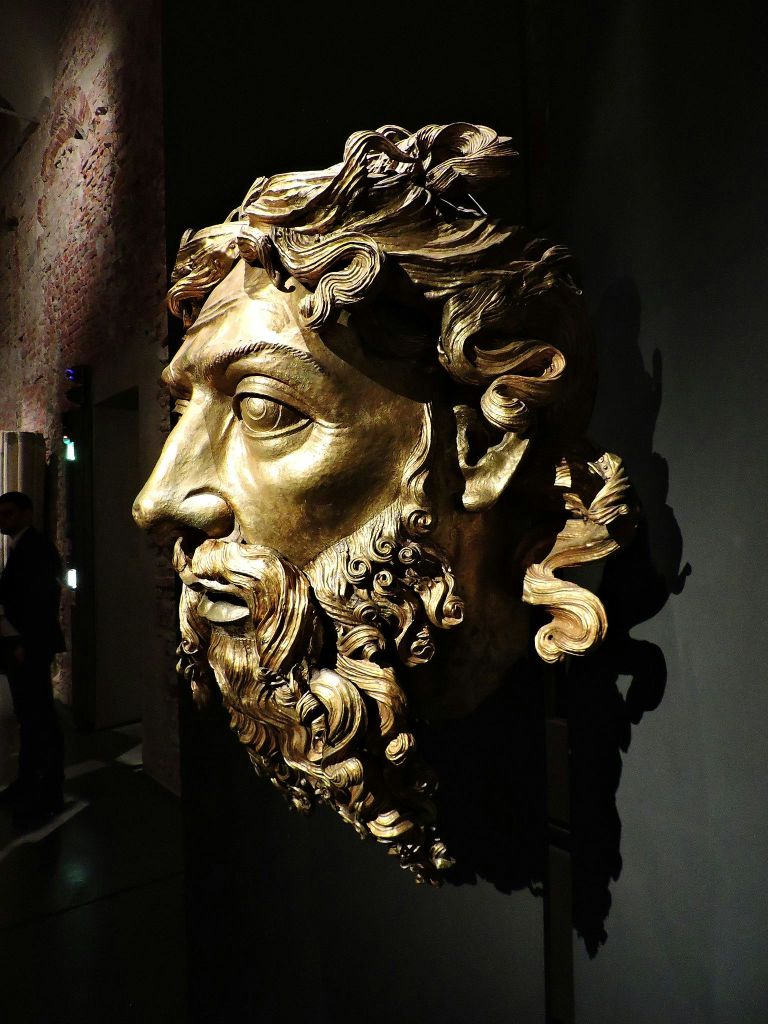 커다랗고 정교하게 조각된 황금빛 얼굴에 곱슬머리와 수염이 어둡게 빛나는 방의 벽에 전시되어 있습니다.