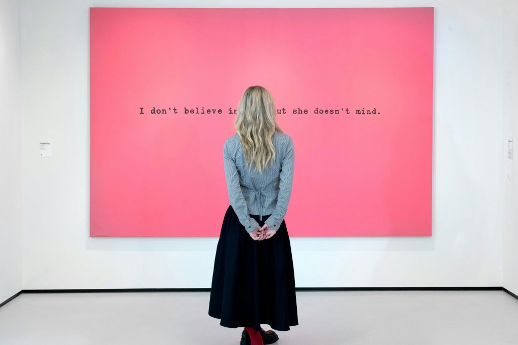 금발의 긴 머리카락을 가진 사람이 벽에 걸린 분홍색 작품을 마주보고 서 있습니다. 이 작품에는 "나는 예술을 믿지 않지만 그녀는 신경 쓰지 않는다" 라는 문구가 적혀 있습니다.
