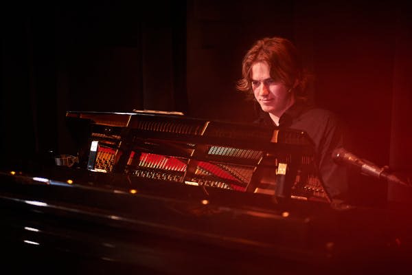 붉은 무대 조명 아래에서 긴 머리카락을 가진 사람이 그랜드 피아노를 연주하고 근처에 마이크가 놓여 있습니다.