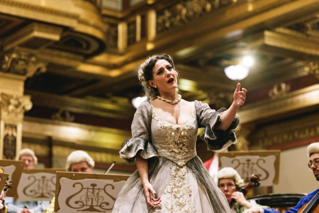 화려하고 역사적인 드레스를 입은 한 여성이 금색으로 장식된 웅장한 콘서트홀에서 열정적으로 노래하고 있으며, 뒤에는 오케스트라 음악가들이 있습니다.