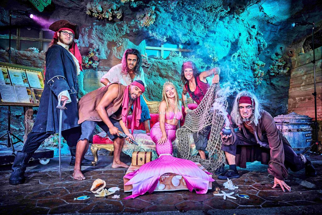 해적으로 옷을 입은 한 무리의 사람들과 인어가 보물 상자와 조개껍질 같은 소품이 있는 수중 테마 세트를 입고 포즈를 취하고 있습니다.