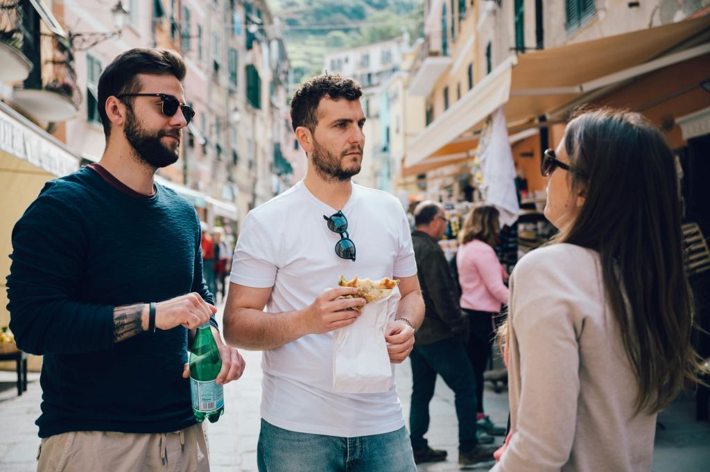 캐주얼한 옷을 입은 두 남성이 번잡한 거리에 서 있습니다. 한 명은 음료를 들고, 다른 한 명은 음식을 들고 있습니다. 배경에는 사람과 건물이 있습니다.