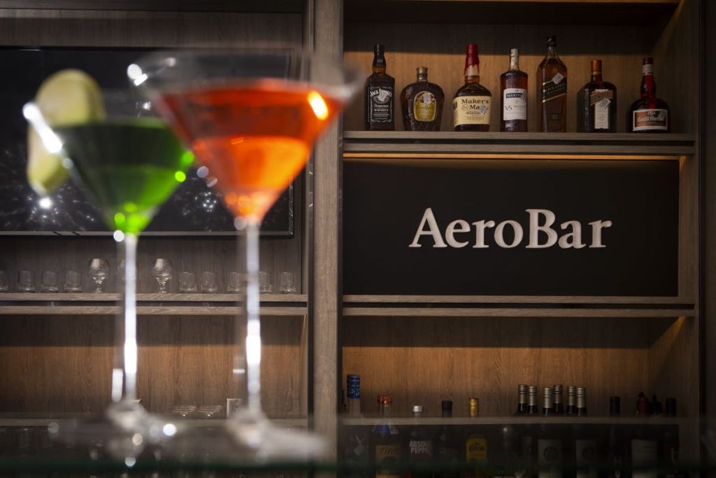 초록색과 주황색 음료가 담긴 두 개의 마티니 잔, 그 뒤에는 술병이 놓인 선반과 "AeroBar" 라고 적힌 간판이 있습니다.